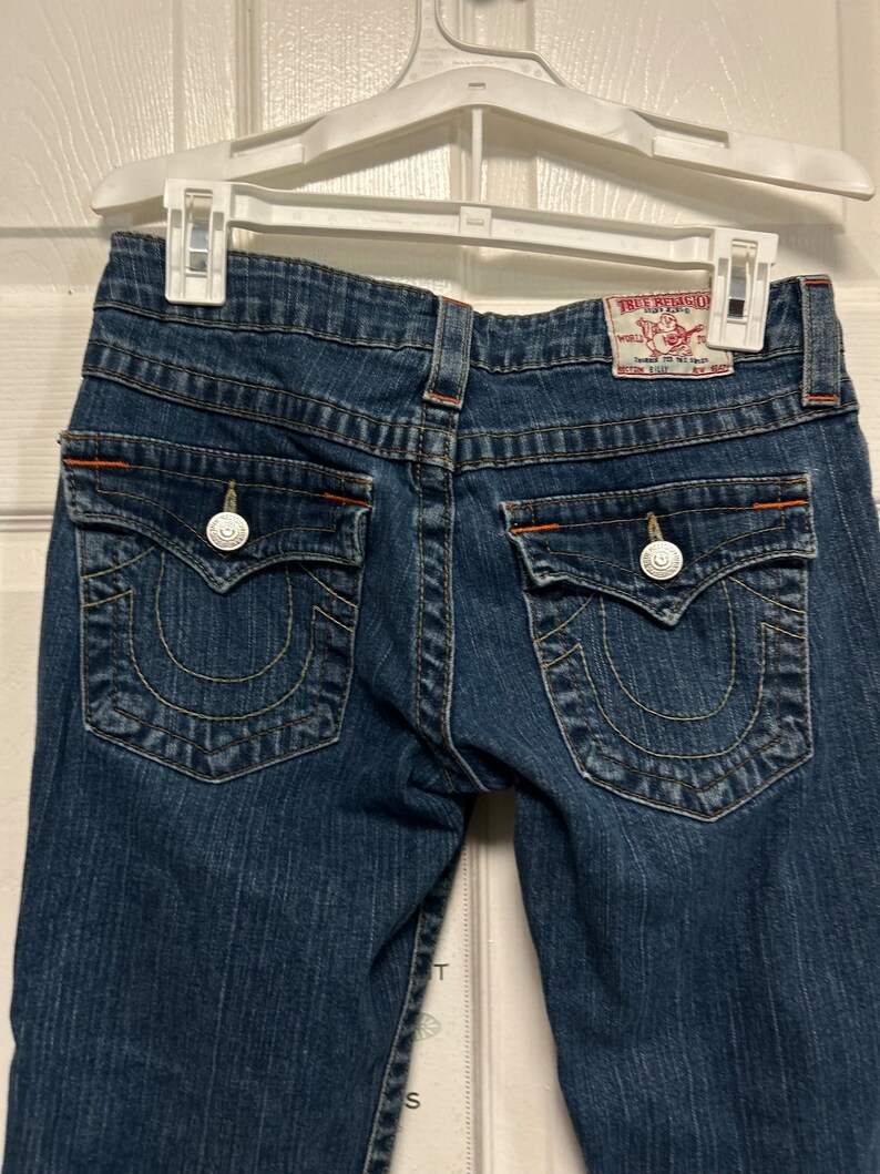 May include: Dark blue denim jeans with decorative stitching and orange accents. The jeans have two back pockets with silver button closures and a True Religion brand label.