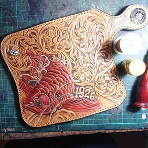 Könnte beinhalten: Handgefertigte Ledergeldbörse mit einem roten Fischdesign, dem Jahr "1926" und floralen Mustern. Die Geldbörse hat einen hellbraunen Hintergrund mit detaillierten Schnitzereien und einem dekorativen Rand. Die Geldbörse liegt auf einer grünen Schneidematte.