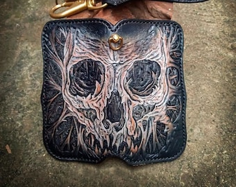 May include: A black leather wallet with a skull design embossed on the front. The skull is white and has a detailed design. The wallet has a strap and a metal clasp.