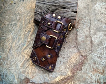 Op de afbeelding: Een bruin leren polsbandje met metalen accenten en gespen. Het bandje heeft een verweerde look en is versierd met klinknagels en stiksels.