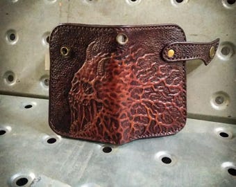 Könnte beinhalten: Eine braune Lederbörse mit einem geprägten Totenkopfdesign auf der Vorderseite. Die Börse hat eine Klappe mit einem Druckknopfverschluss und einen kleinen Riemen mit einer Metallschnalle.