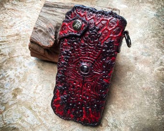 Könnte beinhalten: Eine handgefertigte, rot-schwarze Ledergeldbörse mit einem strukturierten, organischen Design. Die Geldbörse hat einen zentralen, dunkel gefärbten Knopf und einen Metallring zur Befestigung. Die Lederarbeit ist detailliert und aufwendig.