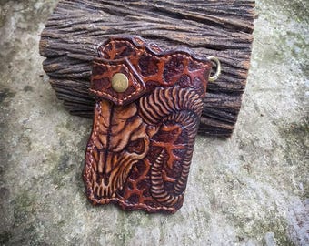May include: A brown leather key chain with a tooled design of a skull with horns. The key chain has a brass snap closure and a brass loop for attaching keys.
