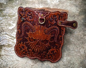 May include: A brown leather wallet with a skull and floral design. The wallet has a metal ring and a strap with a button.