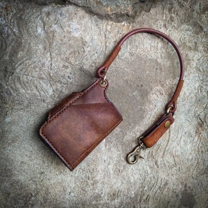 May include: A brown leather wallet with a strap and a metal clasp. The wallet is hanging from a brown leather strap.