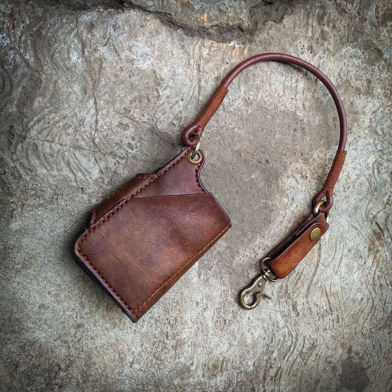 May include: A brown leather wallet with a strap and a metal clasp. The wallet is hanging from a brown leather strap.