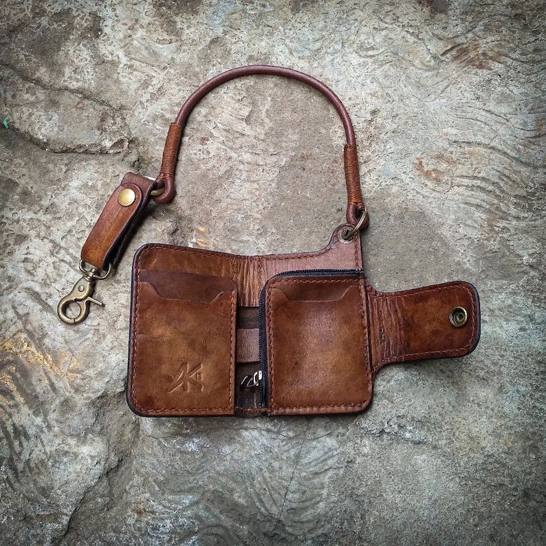 May include: A brown leather wallet with a chain attached. The wallet has multiple compartments and a flap closure. The wallet is on a textured gray surface.