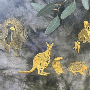 May include: Embroidered fabric featuring Australian animals in gold thread on a tie-dye background. The design includes an emu, koala, kangaroo, wombat, echidna, and eucalyptus leaves. A spool of gold thread is visible.