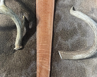 May include: A brown leather strap with a textured pattern and white stitching. The strap is centered in the image, flanked by two pieces of deer antlers. The background is a textured, brown surface.