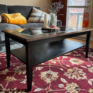 May include: A black coffee table with a glass top and a lower shelf. The table has a wood frame and is sitting on a red and cream patterned rug.