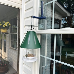 May include: A green, cone-shaped bird feeder with a clear seed container and a white base is mounted outside a window. The feeder is suspended from a blue metal arm attached to the house siding. The window reflects the surrounding trees.
