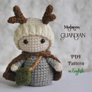 May include: A crocheted doll with a mushroom-themed design. The doll has a cream-colored hat with brown antlers, a gray body, and a brown cape. It also has a green bag and the text "Mushroom Guardian PDF Pattern in English".