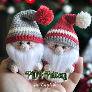 Puede incluir: Dos gnomos de crochet con gorros a rayas rojas y blancas y barbas blancas. Los gnomos se toman de la mano y están sentados en una mano. El texto "PDF Pattern in English" es visible debajo de los gnomos.