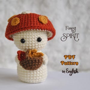 May include: A crocheted mushroom figure with a red cap adorned with yellow spots. The figure has a cream-colored body, black eyes, and holds a brown and orange acorn. Text on the image reads "Forest Spirit" and "PDF Pattern in English."