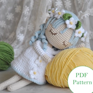 May include: A crocheted doll with blue hair and a white dress, wearing a flower crown. The doll is lying down with a ball of yellow yarn beside it. The text "PDF Pattern" is visible in the lower right corner of the image.