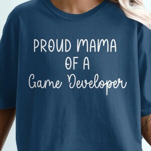 May include: A woman wearing a dark blue t-shirt with the text "Proud Mama of a Game Developer" in white lettering. She is sitting on a white chair with her legs crossed.