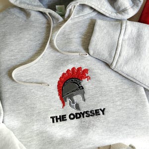 May include: A light gray hooded sweatshirt featuring an embroidered design of a silver and red Spartan-style helmet with a red crest. Below the helmet, the text THE ODYSSEY is embroidered in black thread. Two spools of red and black thread are visible.