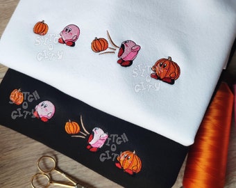 May include: A white sweatshirt and a black sweatshirt, each featuring an embroidered sequence of a pink character interacting with orange pumpkins. The design shows the character approaching, inhaling, and then appearing inside a pumpkin. The text Stitch City is visible.