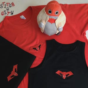 Può includere: Una t-shirt rossa, una t-shirt nera e una canotta nera, ognuna con un logo ricamato a forma di pipistrello rosso con il volto di un personaggio. Un peluche con testa rossa e corpo color crema è appoggiato sulla maglietta rossa. Il testo STITCH O CITY è visibile.