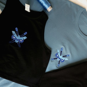 Peut inclure: Une collection de vêtements comprenant un t-shirt bleu clair et deux hauts noirs, dont un débardeur court. Chaque vêtement présente un logo brodé en forme de masque bleu avec des outils croisés. Le texte Stitch City est visible dans le coin.