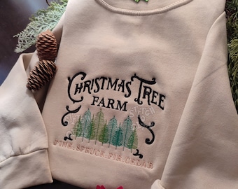 Può includere: Una felpa beige con un ricamo che dice "Christmas Tree Farm" con una fila di alberi verdi sotto e il testo "Pine, Spruce, Fir, Cedar".