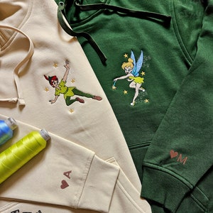 Pode incluir: Duas sweatshirts com capuz, uma bege e outra verde, com desenhos bordados. A sweatshirt bege tem um desenho animado do Peter Pan voando com um design de estrela e o texto "Stitch City". A sweatshirt verde tem um desenho animado da Sininho voando com um design de estrela. Ambas as sweatshirts têm um pequeno coração com uma letra bordada na manga.