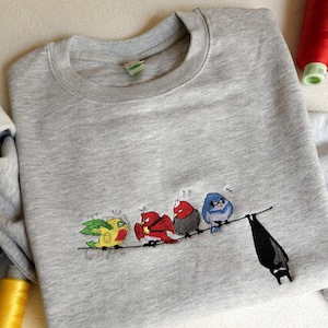 Peut inclure: Un sweat-shirt gris clair à col rond orné d'une broderie représentant quatre oiseaux colorés perchés sur un fil et une chauve-souris noire suspendue à l'envers au même fil. Les oiseaux sont jaunes, rouges, gris et bleus avec des expressions.