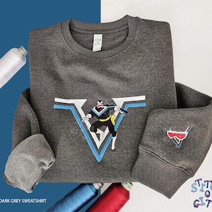 Op de afbeelding: Donkergrijze sweatshirt met een superheldenafbeelding. Het ontwerp toont een superheld in zwart, geel en wit, binnen een blauw en wit V-vormig embleem. Een kleiner embleem staat op de mouw. De tekst "Stitch O City" staat rechtsonder.