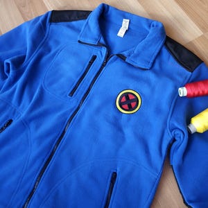 Op de afbeelding: Een felblauw fleece jack met ritssluiting, zwarte schouderstukken en een ronde badge op de borst met een rood en zwart X-symbool met een gele rand. Het jack is voorzien van een verticale borstzak met rits en twee zakken aan de onderzijde.