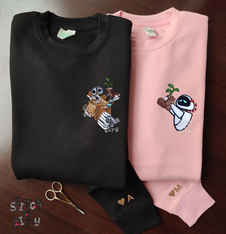 May include: Two black and pink sweatshirts with embroidered images of a robot holding a plant. The black sweatshirt has the text "Stitch City" embroidered on the front. The pink sweatshirt has the letter "M" embroidered on the cuff.