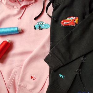 May include: Two hooded sweatshirts, one pink and one black, with embroidered cartoon cars on the front. The pink sweatshirt has a red heart with the letter "D" embroidered on the left sleeve. The black sweatshirt has a blue heart with the letter "E" embroidered on the left sleeve. The text "Stitch City" is embroidered on the bottom right corner of the black sweatshirt.