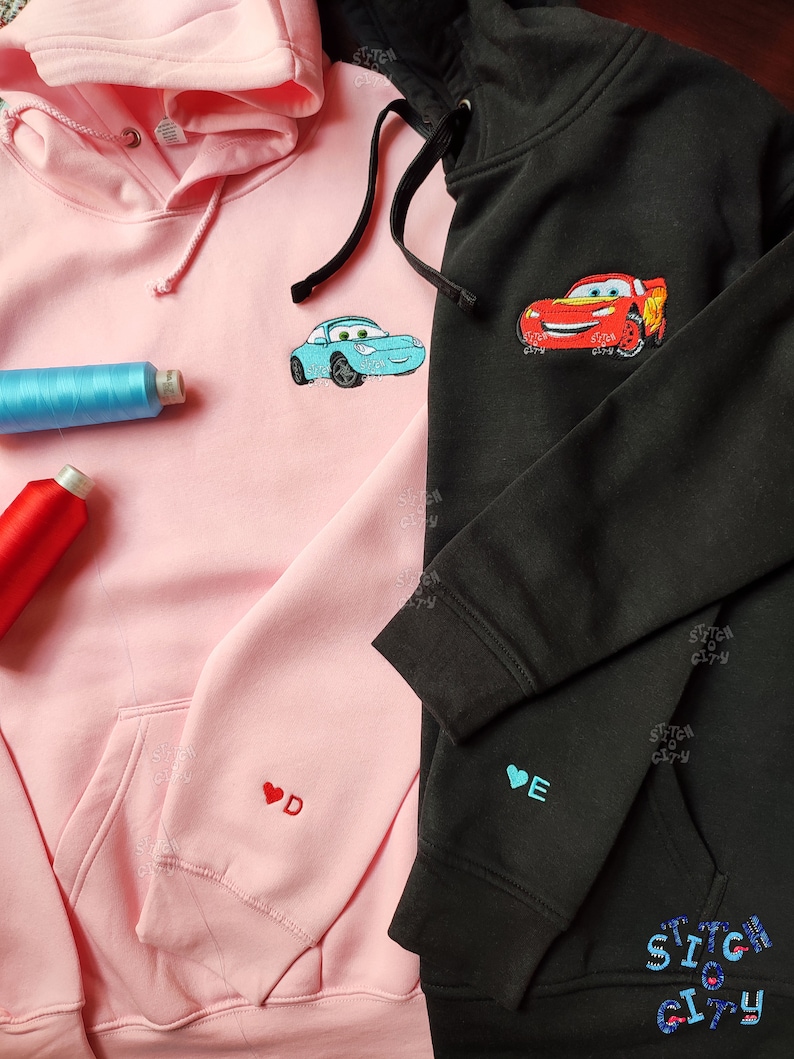 May include: Two hooded sweatshirts, one pink and one black, with embroidered cartoon cars on the front. The pink sweatshirt has a red heart with the letter "D" embroidered on the left sleeve. The black sweatshirt has a blue heart with the letter "E" embroidered on the left sleeve. The text "Stitch City" is embroidered on the bottom right corner of the black sweatshirt.