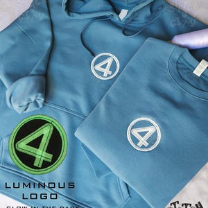 Op de afbeelding: Twee blauwe sweatshirts, een hoodie en een crew neck, met een wit en blauw logo met het nummer 4. De hoodie heeft een lichtgevend groen logo dat oplicht in het donker. De afbeelding bevat de tekst "Luminous Logo" en "Glow in the Dark."