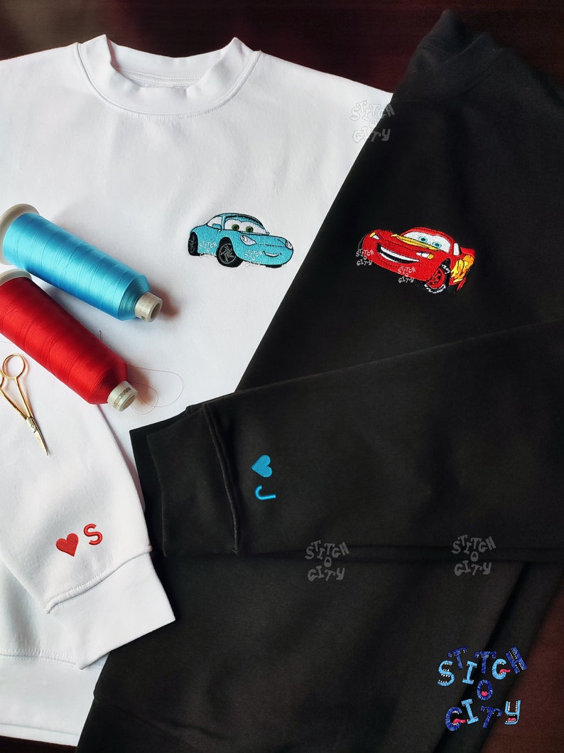 May include: Two sweatshirts with embroidered images of cartoon cars. The white sweatshirt has a blue car with a red heart on the sleeve. The black sweatshirt has a red car with a blue heart on the sleeve. Both sweatshirts have the text "Stitch City" on the sleeve.