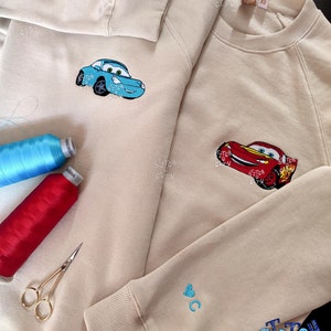 May include: Two beige sweatshirts with embroidered designs. One sweatshirt has a blue car with a red heart on the sleeve. The other sweatshirt has a red car and a blue heart on the sleeve. The text "Stitch City" is embroidered on the bottom of the sweatshirt.
