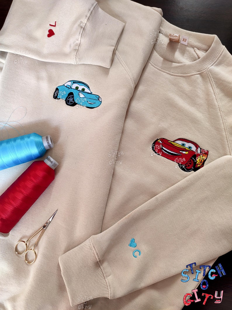May include: Two beige sweatshirts with embroidered designs. One sweatshirt has a blue car with a red heart on the sleeve. The other sweatshirt has a red car and a blue heart on the sleeve. The text "Stitch City" is embroidered on the bottom of the sweatshirt.