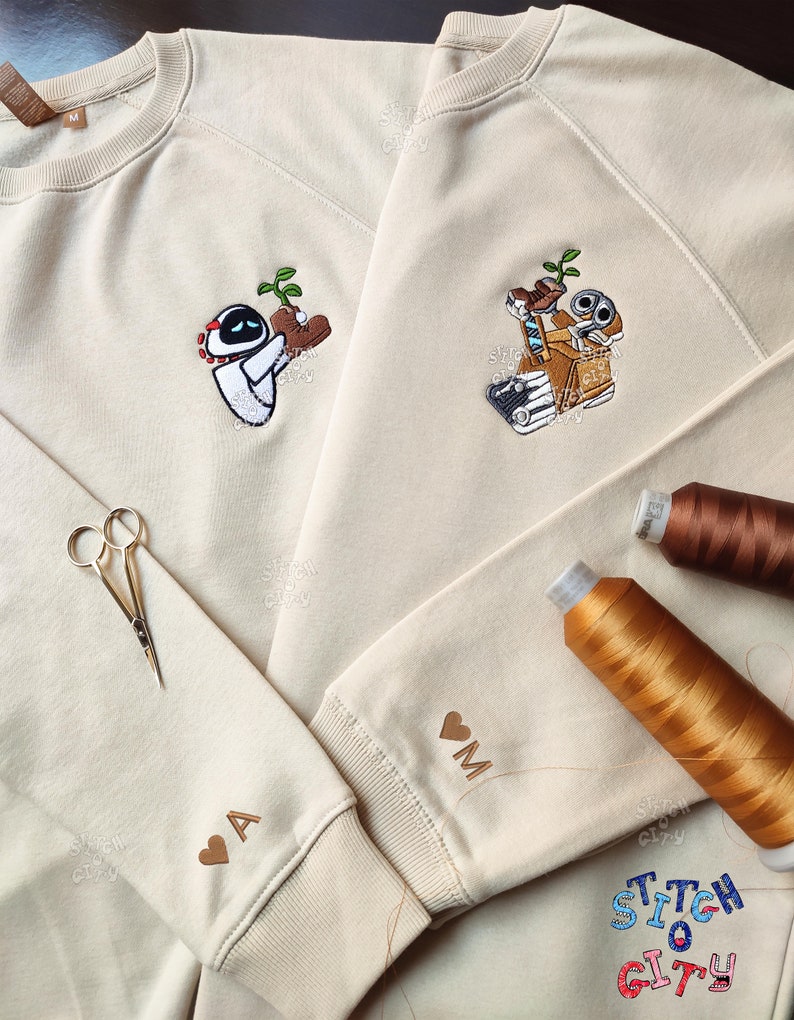 May include: Two beige sweatshirts with embroidered images of Wall-E and Eve from the animated film Wall-E. The sweatshirts have the letters "A" and "M" embroidered on the sleeves. The text "Stitch City" is embroidered on the bottom right corner of one of the sweatshirts.