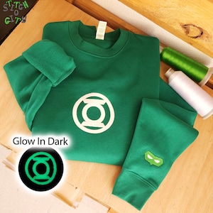 May include: A green crewneck sweatshirt featuring a white circular Green Lantern logo on the chest and a small embroidered green mask on the sleeve. The text Glow In Dark is printed near a circular graphic showing the logo glowing in green.