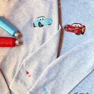 May include: Two gray sweatshirts with embroidered Cars characters. One sweatshirt has a blue Lightning McQueen and the other has a red Mater. The sleeves have embroidered hearts with the initials J and K.