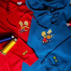 Könnte beinhalten: Rote und blaue Kapuzenpullover mit gestickten Cartoon-Charakteren. Die Kapuzenpullover haben Kordelzüge und Vordertaschen. Ein roter Kapuzenpullover hat den Buchstaben "M" und ein grünes Herz auf dem Ärmel gestickt.