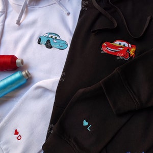 May include: Two hooded sweatshirts with embroidered designs. One is white with a light blue car and a red heart with the letter "D". The other is black with a red car and a blue heart with the letter "L". Both sweatshirts have the text "Stitch City" embroidered on them.