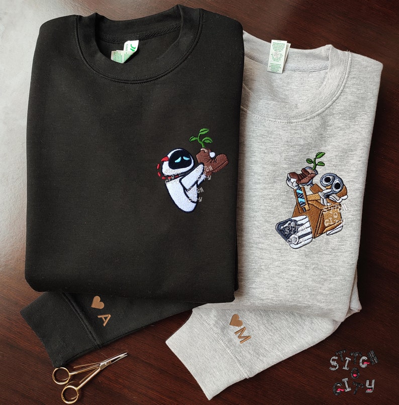 May include: Two black and gray sweatshirts with embroidered images of robots from the movie Wall-E. The black sweatshirt has a robot holding a plant and the gray sweatshirt has a robot holding a box. The sweatshirts have the letters "A" and "M" embroidered on the sleeves.