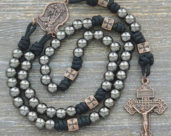 May include: A rosary featuring metallic gray beads strung on black cord, accented with copper-toned metal spacers and a central crucifix. The design includes a detailed copper-toned centerpiece depicting a religious figure, with small cross motifs on the spacers.