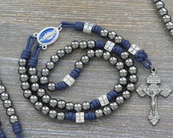 May include: A rosary featuring dark metallic gray beads strung on navy blue cord. The design includes silver-toned square spacers with cross engravings, a blue oval centerpiece depicting a religious figure, and a detailed silver-toned crucifix.