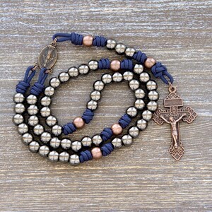 May include: A silver and copper rosary with a navy blue cord. The rosary has a silver crucifix with a copper Jesus figure. The rosary is laid out on a wooden surface.