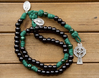 May include: A rosary featuring glossy black beads interspersed with green paracord knots and green marbled beads. The piece includes a silver-toned Celtic cross crucifix and two small oval metal religious medals attached to the corded sections.