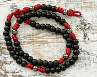 May include: A rosary featuring matte black beads and metallic dark gray spacer beads strung on red cord. The rosary includes a silver-toned crucifix and a circular Saint Benedict medal attached to the cord, arranged in a circular loop pattern.