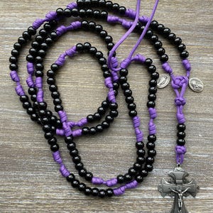 Puede incluir: Un rosario con cuentas negras separadas por nudos de cord&oacute;n morado. El rosario incluye dos peque&ntilde;as medallas met&aacute;licas circulares de tono plateado y un gran crucifijo plateado ornamentado con la figura de Jes&uacute;s y las letras INRI arriba.