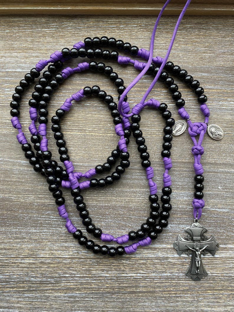 Puede incluir: Un rosario con cuentas negras separadas por nudos de cord&oacute;n morado. El rosario incluye dos peque&ntilde;as medallas met&aacute;licas circulares de tono plateado y un gran crucifijo plateado ornamentado con la figura de Jes&uacute;s y las letras INRI arriba.