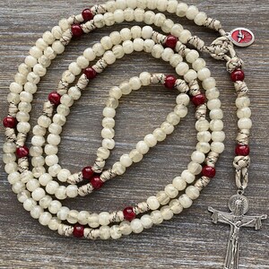 Puede incluir: Un rosario con cuentas transl&uacute;cidas de color blanco roto y cuentas m&aacute;s peque&ntilde;as de color rojo oscuro ensartadas en un cord&oacute;n trenzado. La pieza incluye un crucifijo plateado con la figura de Jes&uacute;s y una peque&ntilde;a medalla ovalada con una paloma roja.